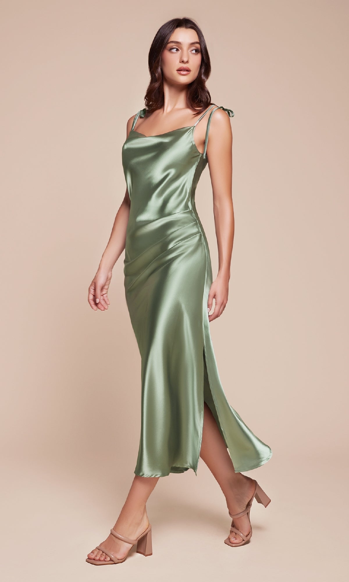 A woman wears the Midi Wedding-Guest Slip Dress BD103 in light green satin with spaghetti straps and a side slit, paired with nude heeled sandals against a beige background. An affordable, elegant choice for wedding guests.